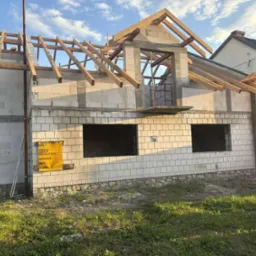 Surowy stan domu z białego bloczka, widoczne drewniane elementy konstrukcji dachu i otwory okienne. Tablica informacyjna budowy na ścianie. Budowa w toku, w słoneczny dzień.