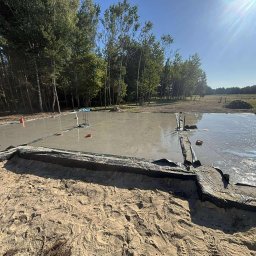 Dan Bud - Zalany fundament pod budowę domu jednorodzinnego. Widoczne szalunki i folia izolacyjna. W tle drzewa i błękitne niebo. Świeży beton wylewany na przygotowany teren.