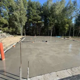 Świeżo wylany fundament pod budowę domu, widoczne pionowe rury i wystające pręty zbrojeniowe. W tle drzewa i częściowo usypany teren.