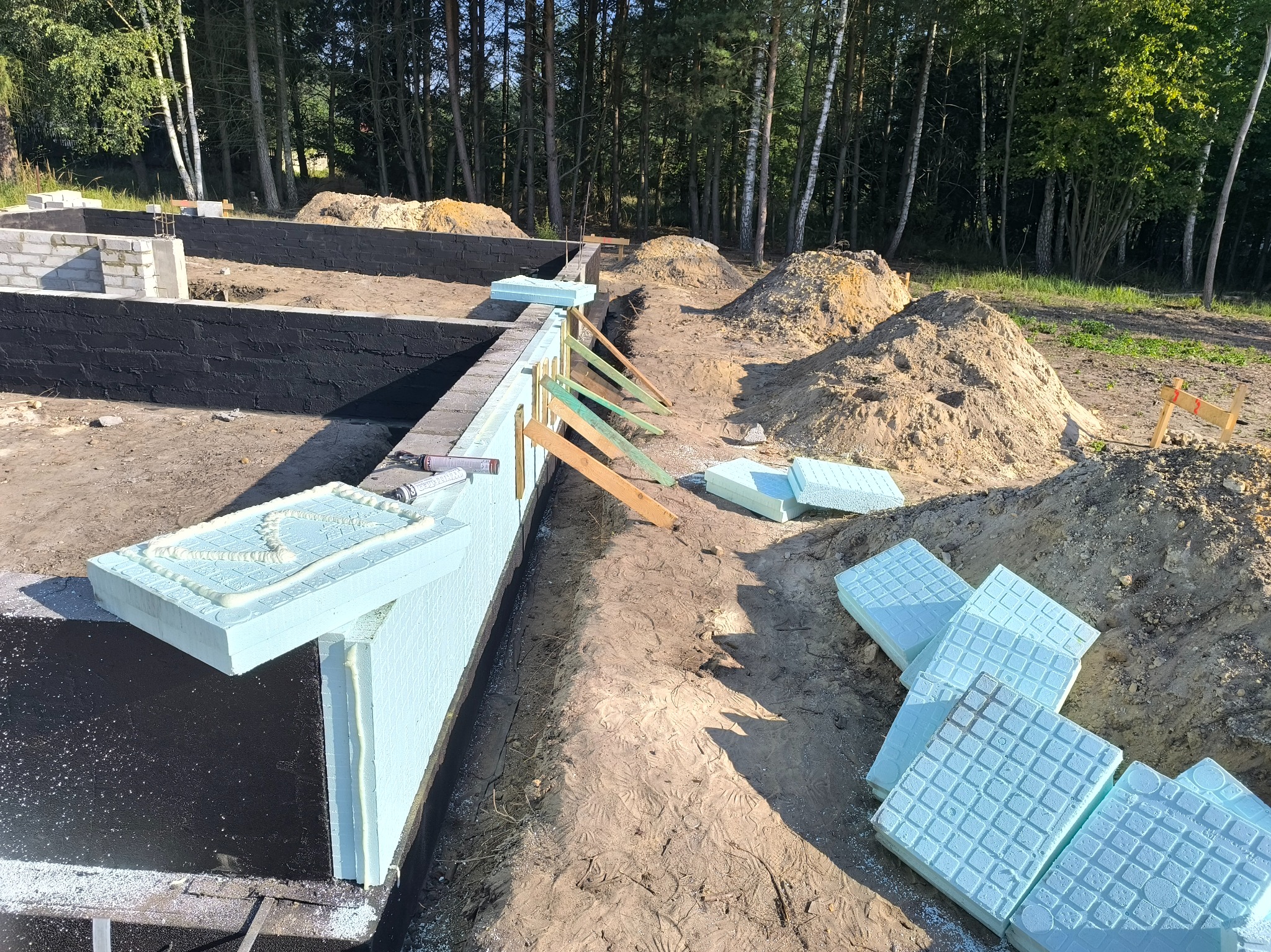 Ocieplenie fundamentów budynku w trakcie budowy, izolacja termiczna ze styropianu, zabezpieczenie hydroizolacyjne, teren budowy z drzewami w tle.