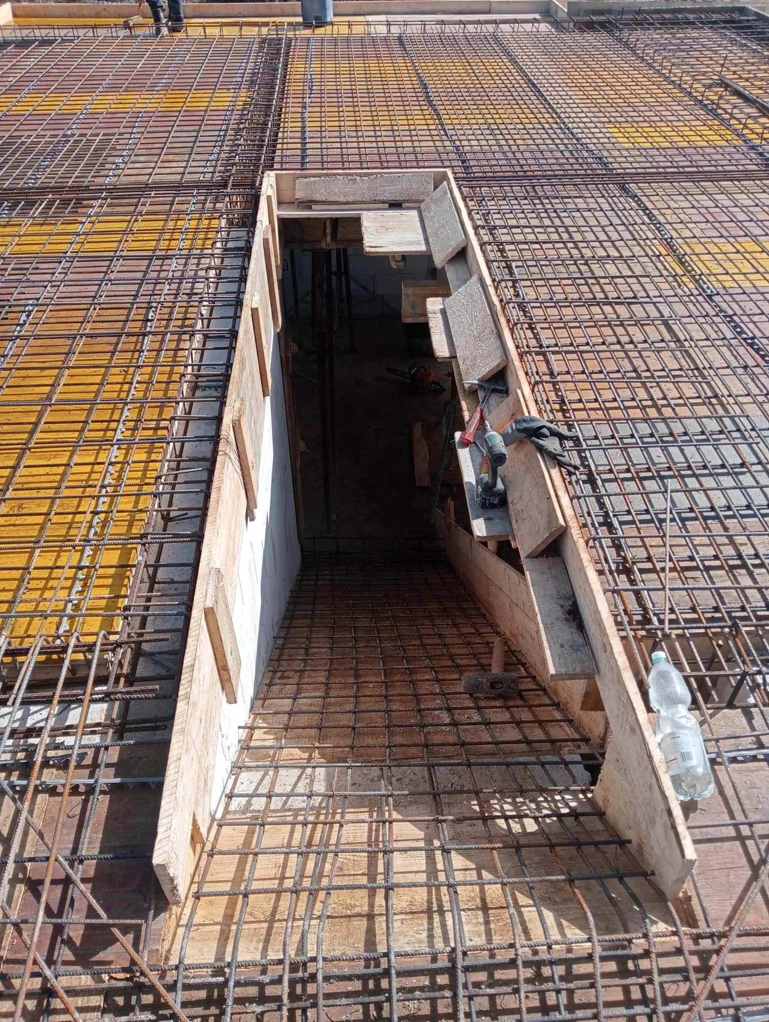 Zbrojenie stropu z otworem na schody, widoczne deski szalunkowe, siatka zbrojeniowa, narzędzia pracy i butelka wody. Prace budowlane w toku, widok z góry.