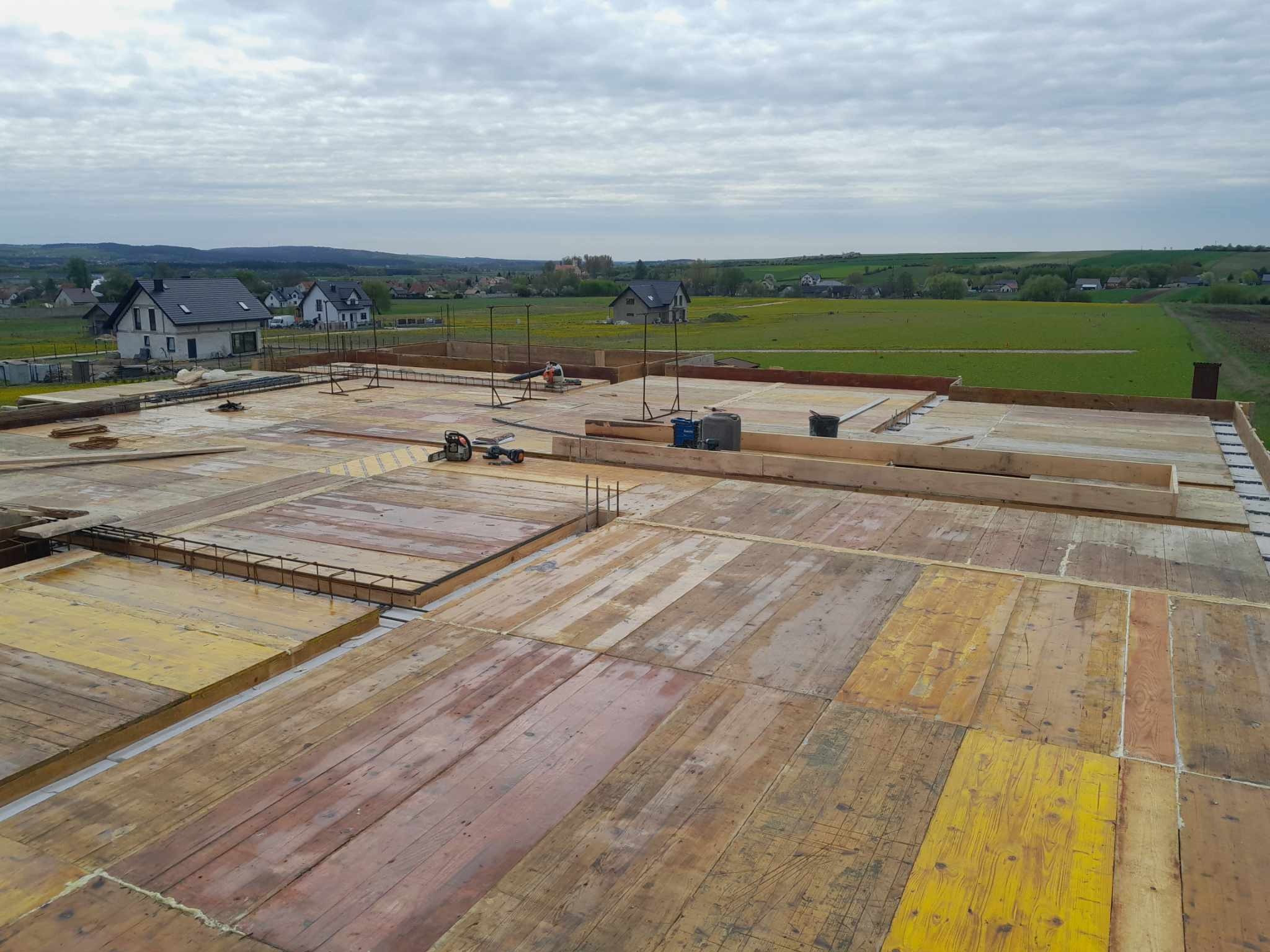 Szeroki kadr na zbrojenie fundamentów domu jednorodzinnego z widokiem na okolicę. Drewniane szalunki, stalowe pręty i narzędzia budowlane na placu budowy.