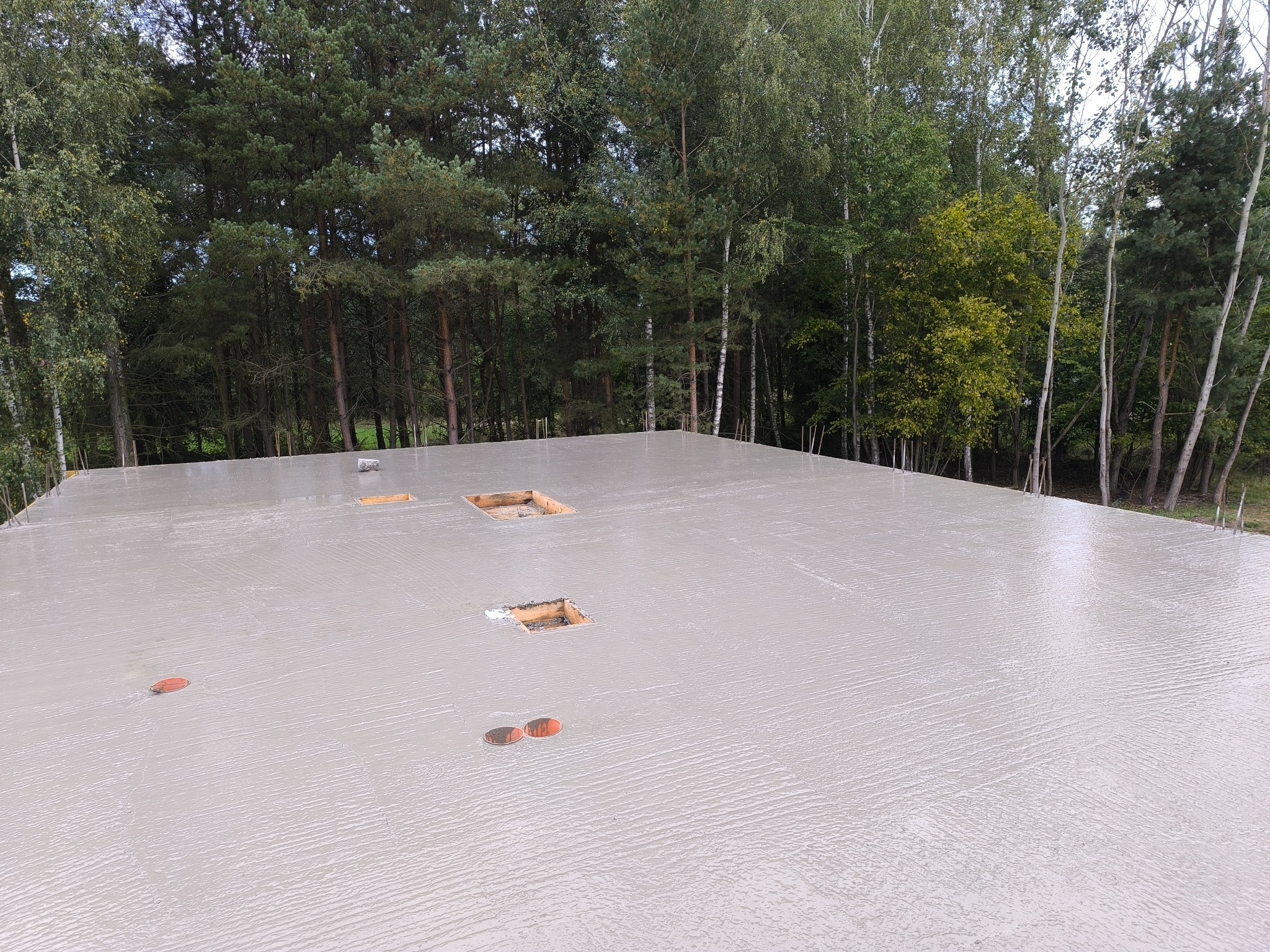 Świeżo wylana, gładka płyta fundamentowa z widocznymi otworami instalacyjnymi, na tle drzew. Widok z góry, podkreślający precyzję wykonania.