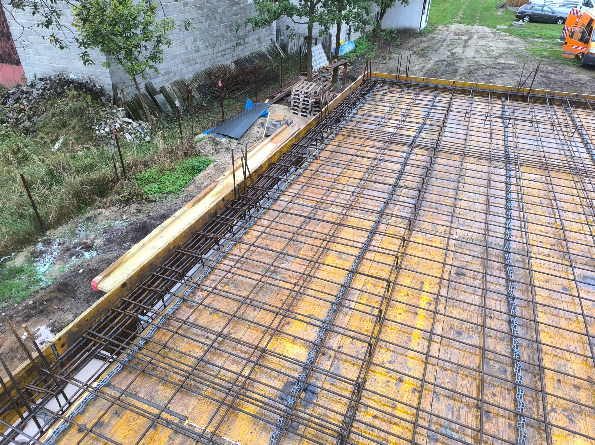 Zbrojenie płyty fundamentowej z użyciem drewnianych szalunków i stalowych prętów. Widok z góry na plac budowy z zielenią w tle.
