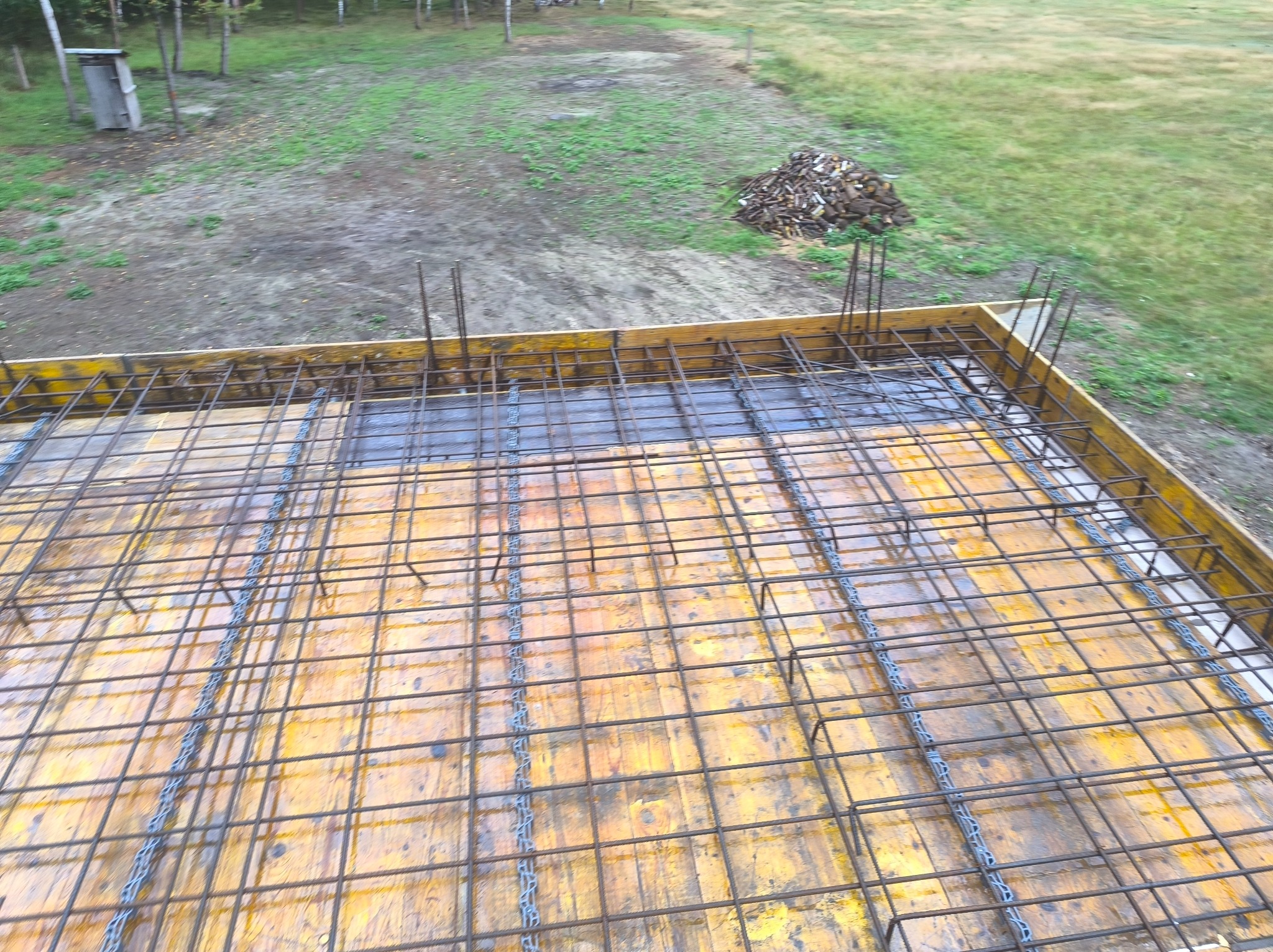 Zbrojenie płyty fundamentowej w szalunku, widok z góry. Na tle zielona trawa i drzewa. Etap budowy fundamentów pod dom.