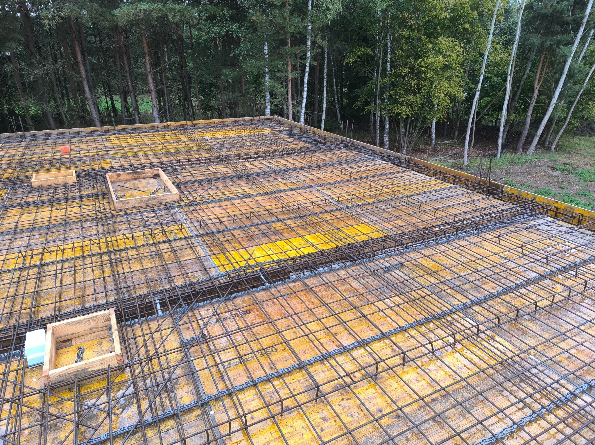 Zbrojenie stropu na żółtym szalunku, widok z góry. Siatka z prętów stalowych, drewniane skrzynki na tle zieleni drzew. Etap przygotowania do wylewania betonu.