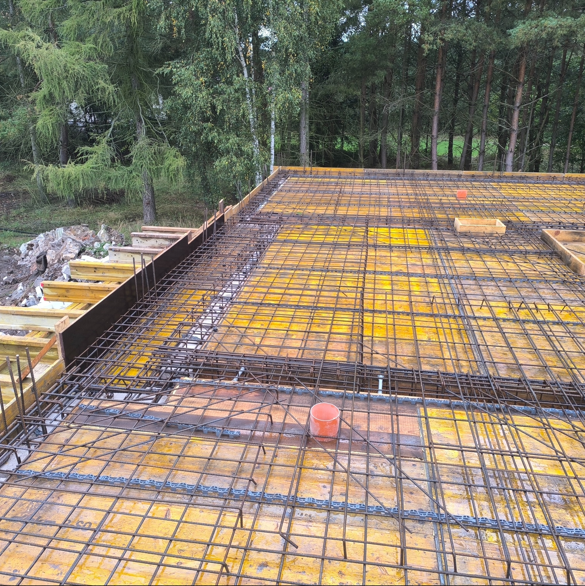 Zbrojenie stropu na deskowaniu z widocznymi elementami instalacji i otaczającym lasem. Układ prętów tworzy gęstą siatkę, przygotowaną do zalania betonem.