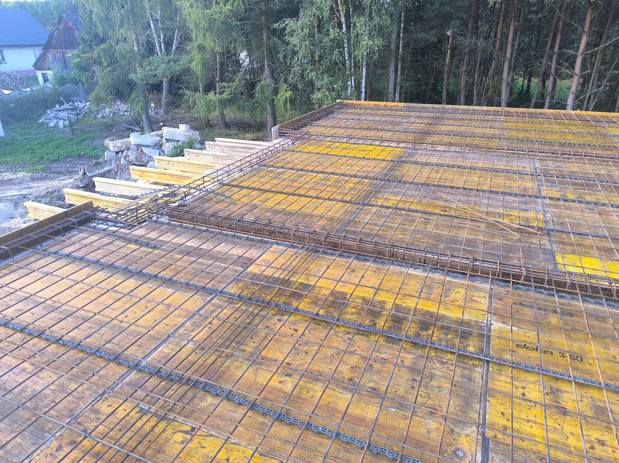 Zbrojenie stropu na deskowaniu z żółtej sklejki, widok z góry. W tle drzewa i fragment budynku. Etap prac przygotowawczych do wylewania betonu.