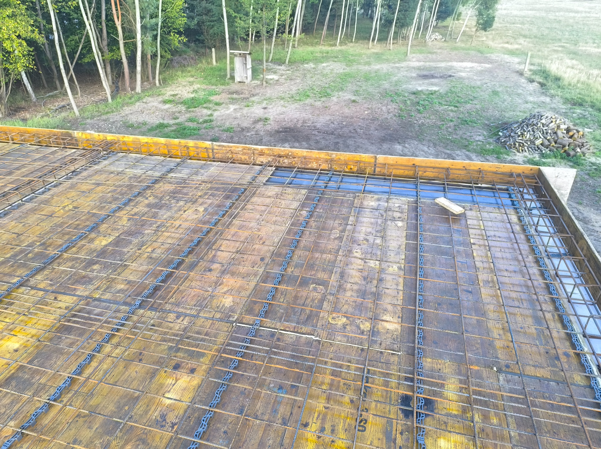 Zbrojenie stropu na deskowaniu, widok z góry. Siatka z prętów stalowych połączona plastikowymi elementami. W tle drzewa i sterta drewna.