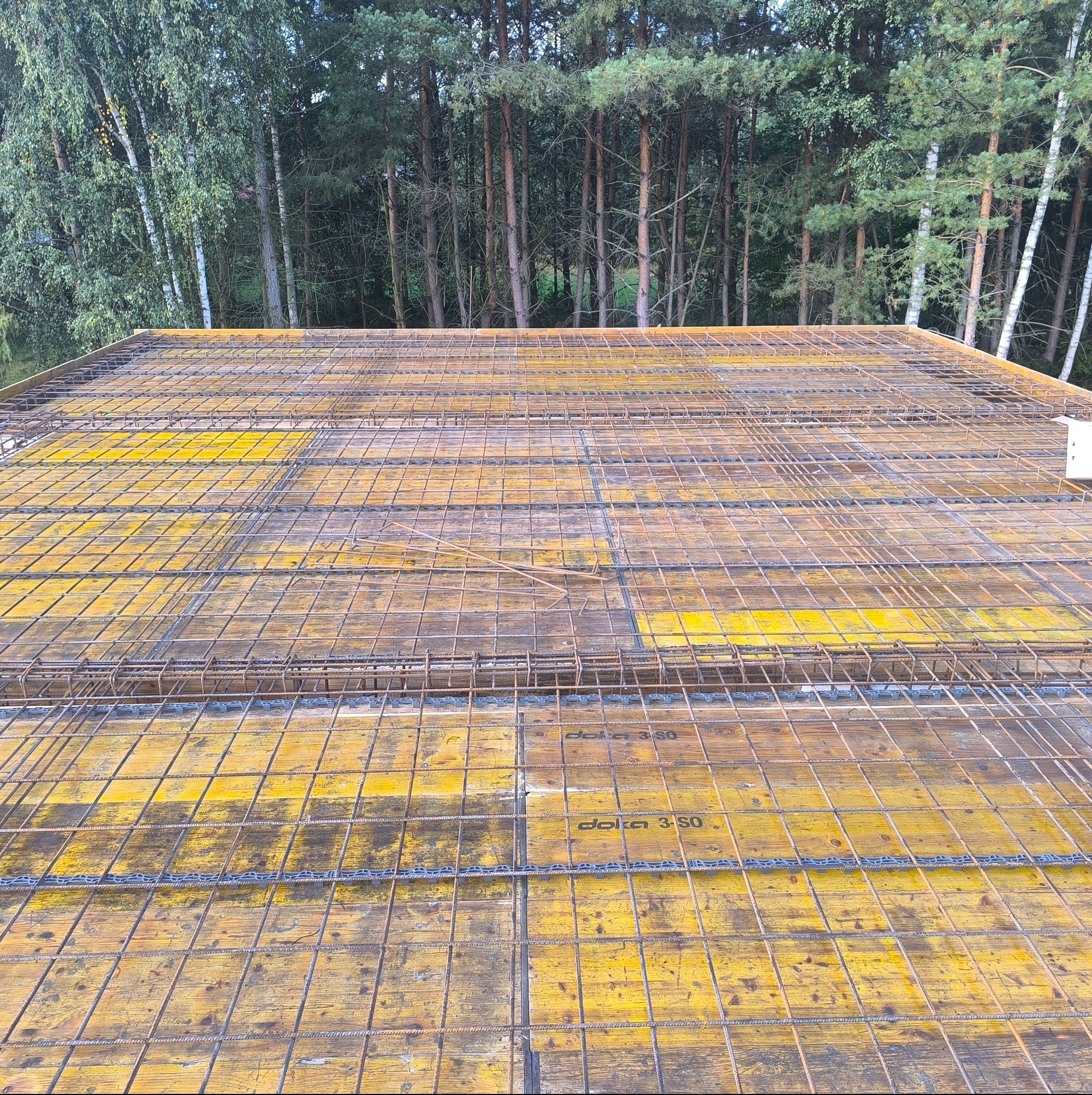 Żółte szalunki Doka 3-SO z ułożonym zbrojeniem na tle lasu. Widok z góry na przygotowany strop budynku w trakcie budowy.
