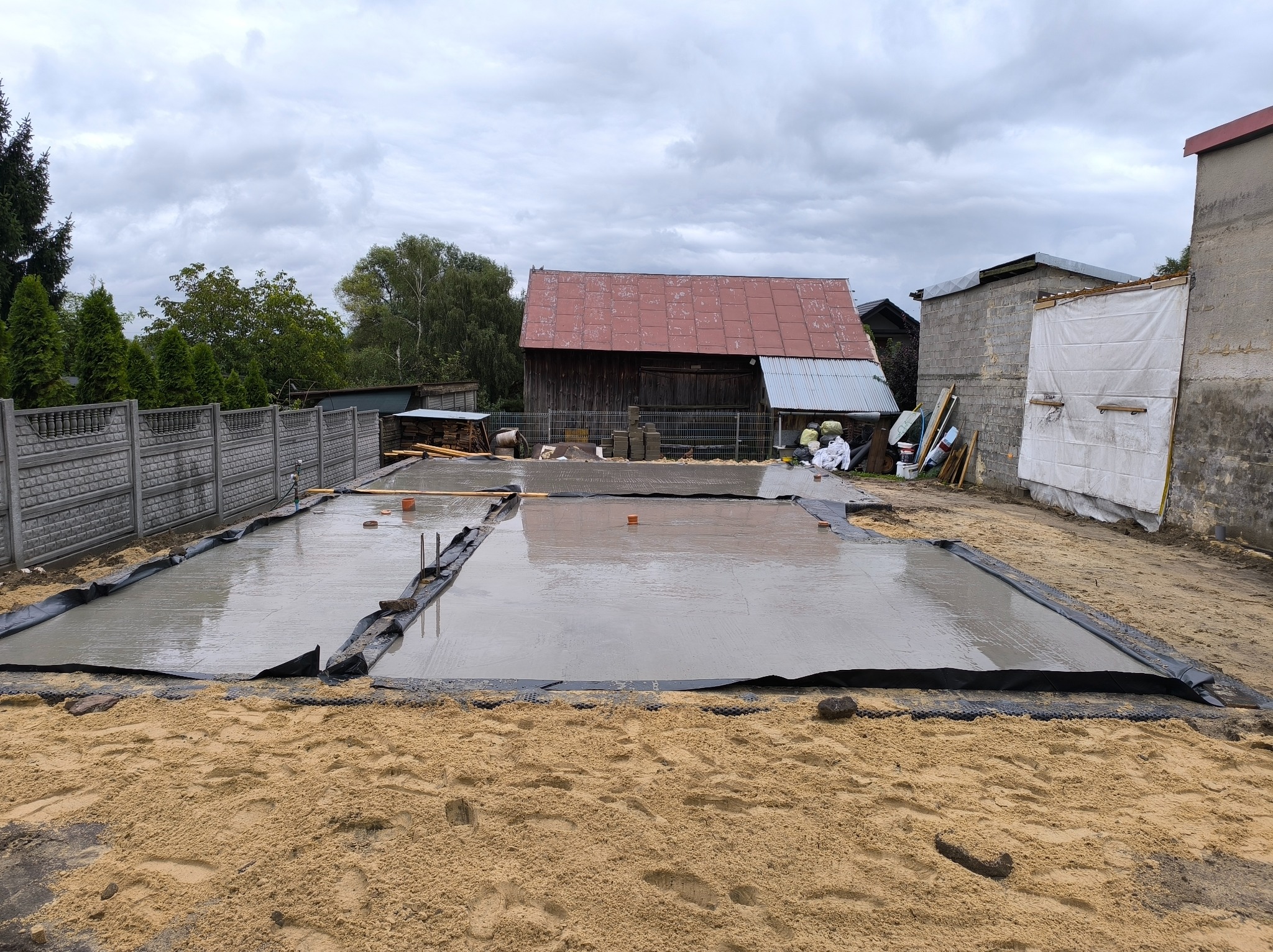 Wylany fundament pod budynek w Sobkowie. Widoczna czarna folia, mokry beton i piasek. W tle budynki gospodarcze i ogrodzenie.