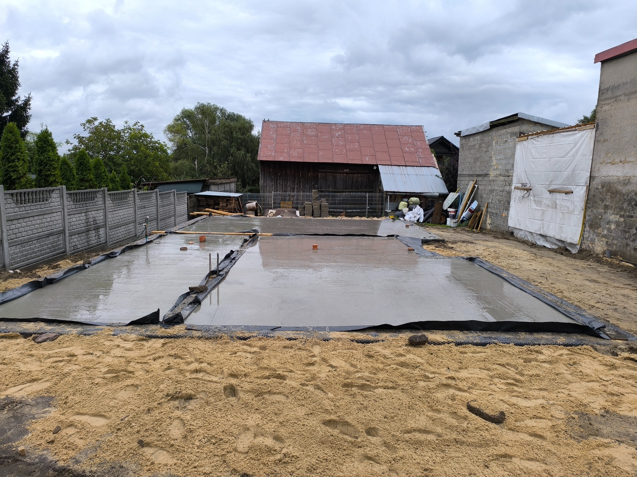 Świeżo wylany fundament na piasku, zabezpieczony czarną folią, z widocznymi elementami szalunku. Tło stanowi zabudowa wiejska i pochmurne niebo.