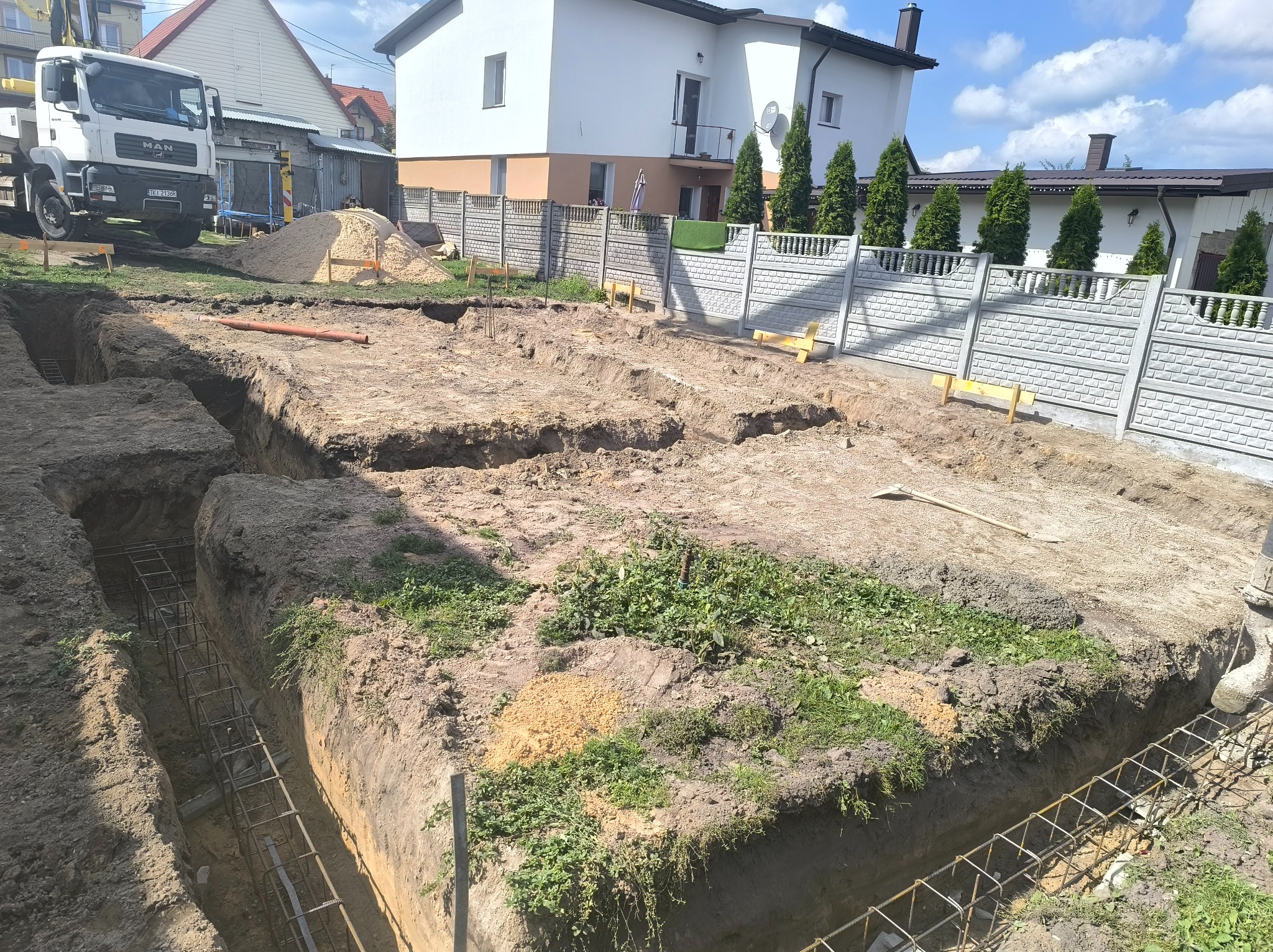 Wykonane wykopy pod fundamenty z widocznym zbrojeniem, na tle domu jednorodzinnego z białymi ścianami i szarym płotem. W tle stoi ciężarówka.