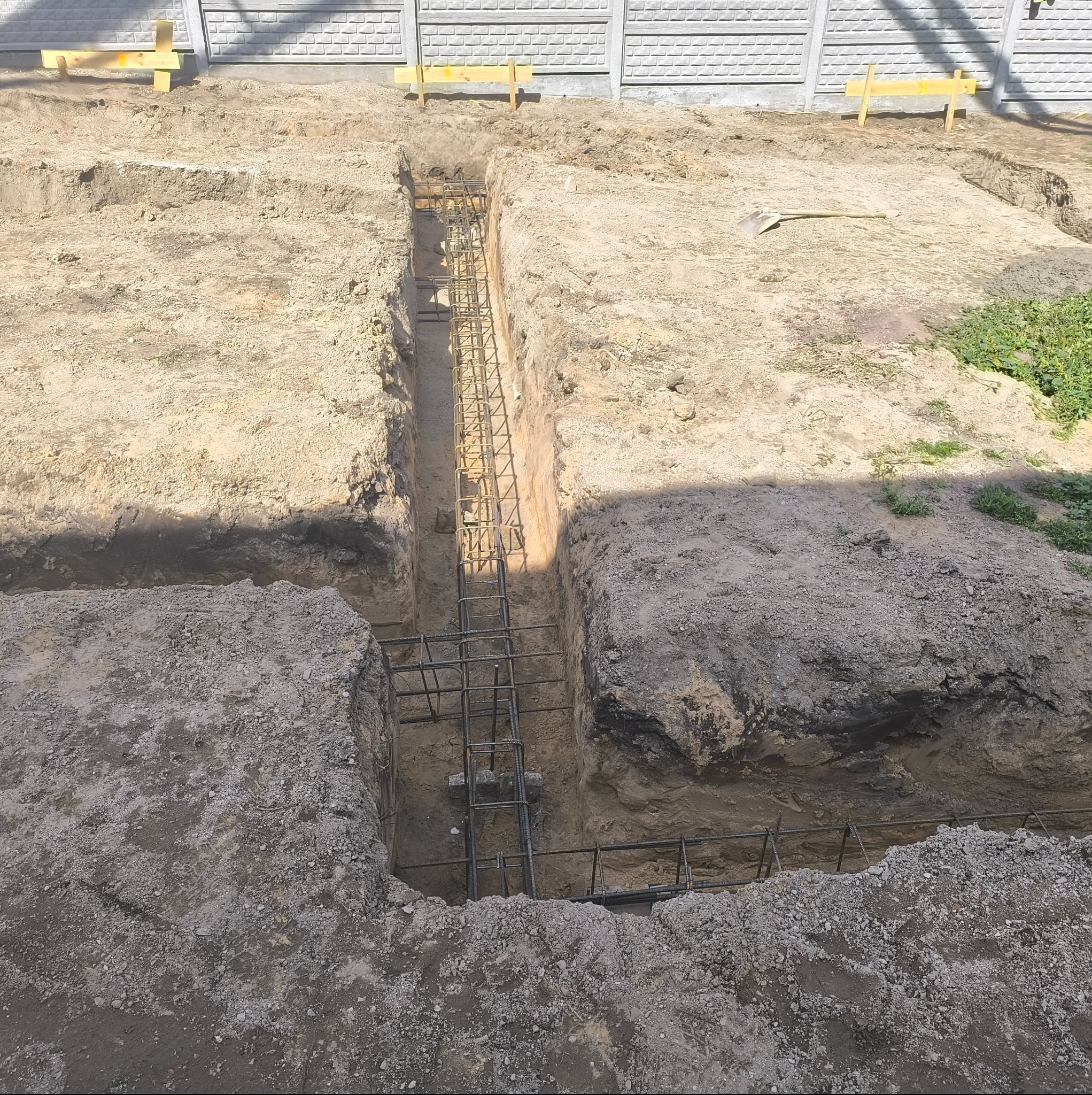Wykop fundamentowy z ułożonym zbrojeniem stalowym, widok z góry. Na pierwszym planie ziemia, w tle fragment ogrodzenia i drewniane elementy szalunku.