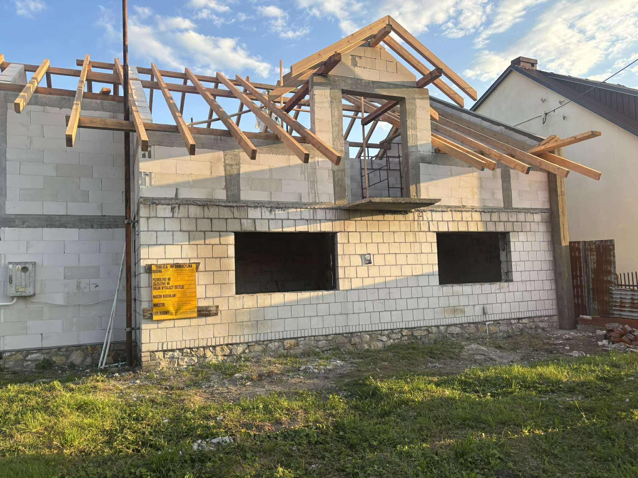 Surowy stan domu z białego bloczka, widoczne drewniane elementy konstrukcji dachu i otwory okienne. Tablica informacyjna budowy na ścianie. Budowa w toku, w słoneczny dzień.