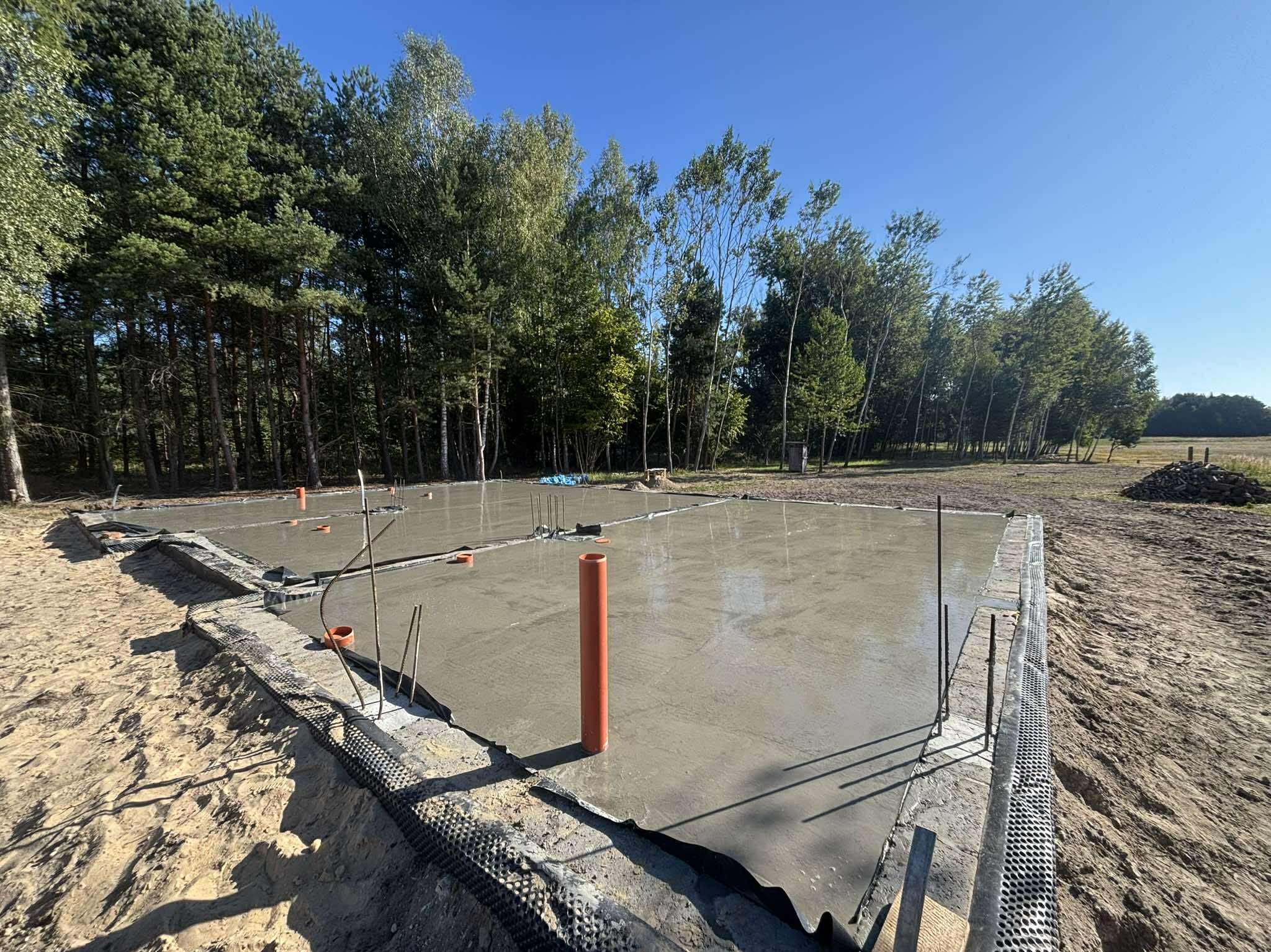 Zalany fundament pod budowę domu jednorodzinnego. Widoczne pionowe rury i folia izolacyjna. W tle drzewa i błękitne niebo.