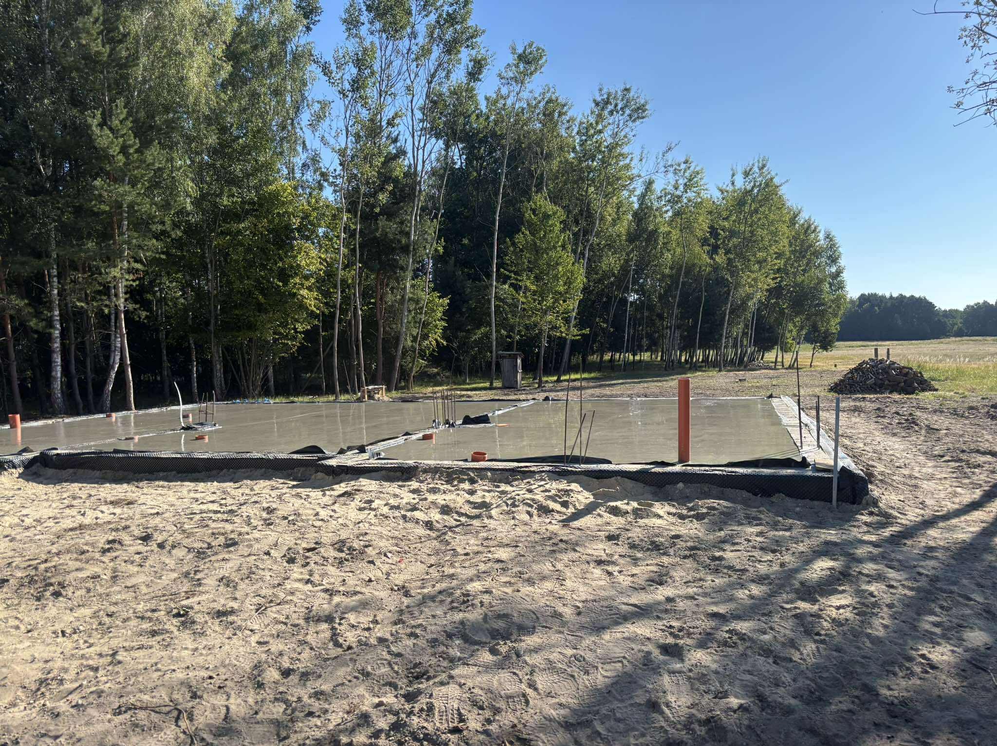 Wylany fundament pod budowę domu, widoczne rury i folia izolacyjna. Tło stanowią drzewa i błękitne niebo. Ujęcie z poziomu gruntu.