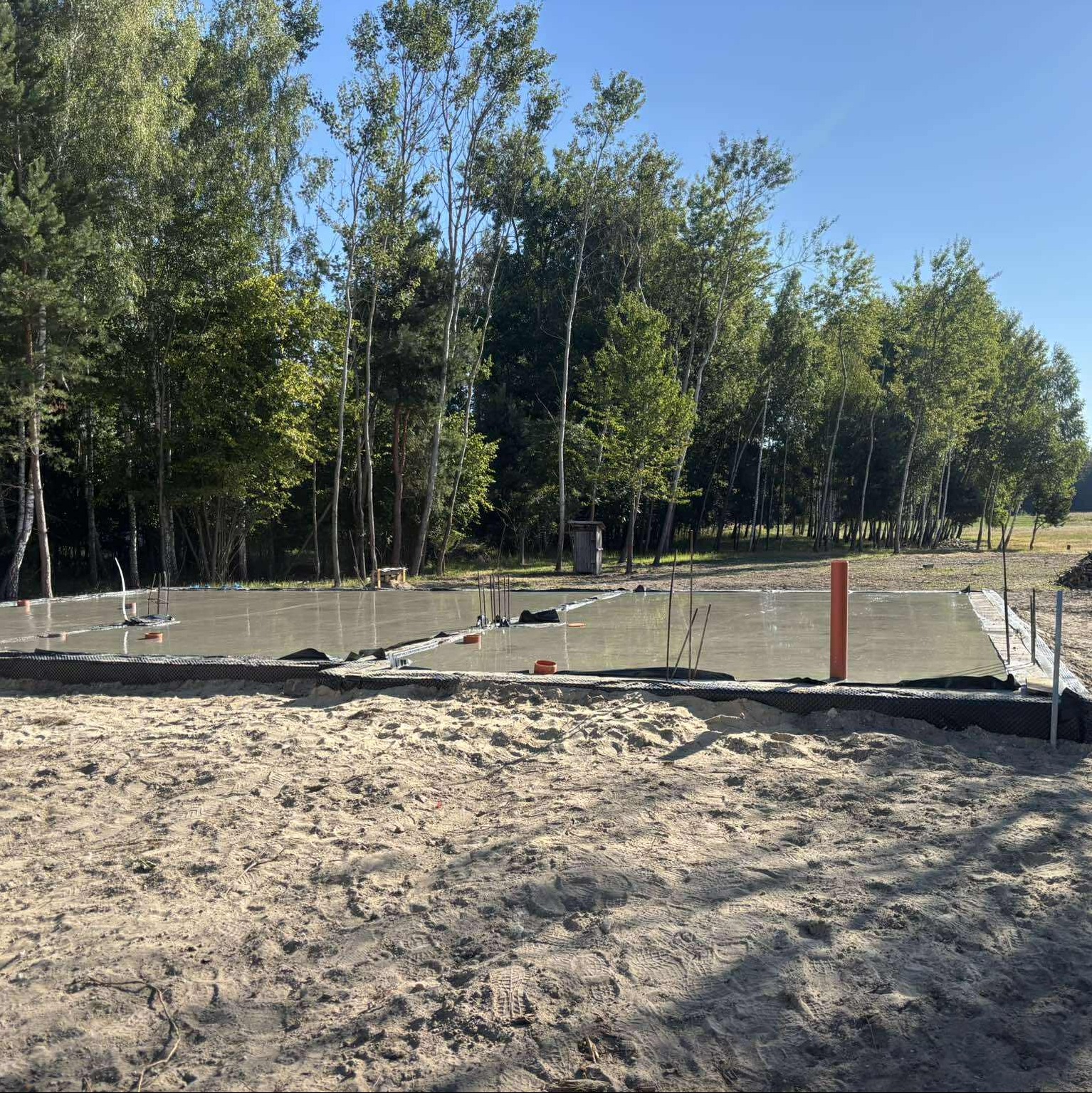 Świeżo wylany fundament pod budowę domu jednorodzinnego, widoczne zbrojenia i szalunki, tło stanowią drzewa i błękitne niebo.