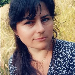 Derevianko Olena - Portret kobiety z ciemnymi włosami na tle łąki w Polkowicach. Ubrana w wzorzystą sukienkę, z delikatnym makijażem i złotym łańcuszkiem. Naturalne światło.