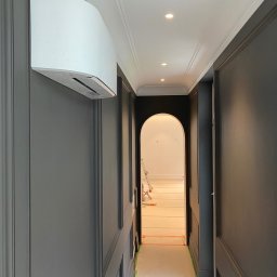 Dita Sp. Z o.o. - Nowoczesny klimatyzator ścienny w szarym korytarzu z dekoracyjnymi panelami i jasnym oświetleniem punktowym. Widok na jasne pomieszczenie przez łukowe przejście.