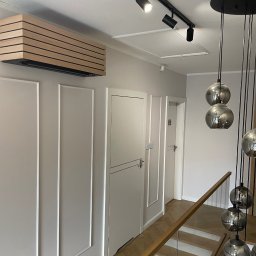 Dita Sp. Z o.o. - Nowoczesny klimatyzator z drewnianą obudową, zamontowany na ścianie w minimalistycznym wnętrzu z oświetleniem szynowym i szklaną balustradą schodów.