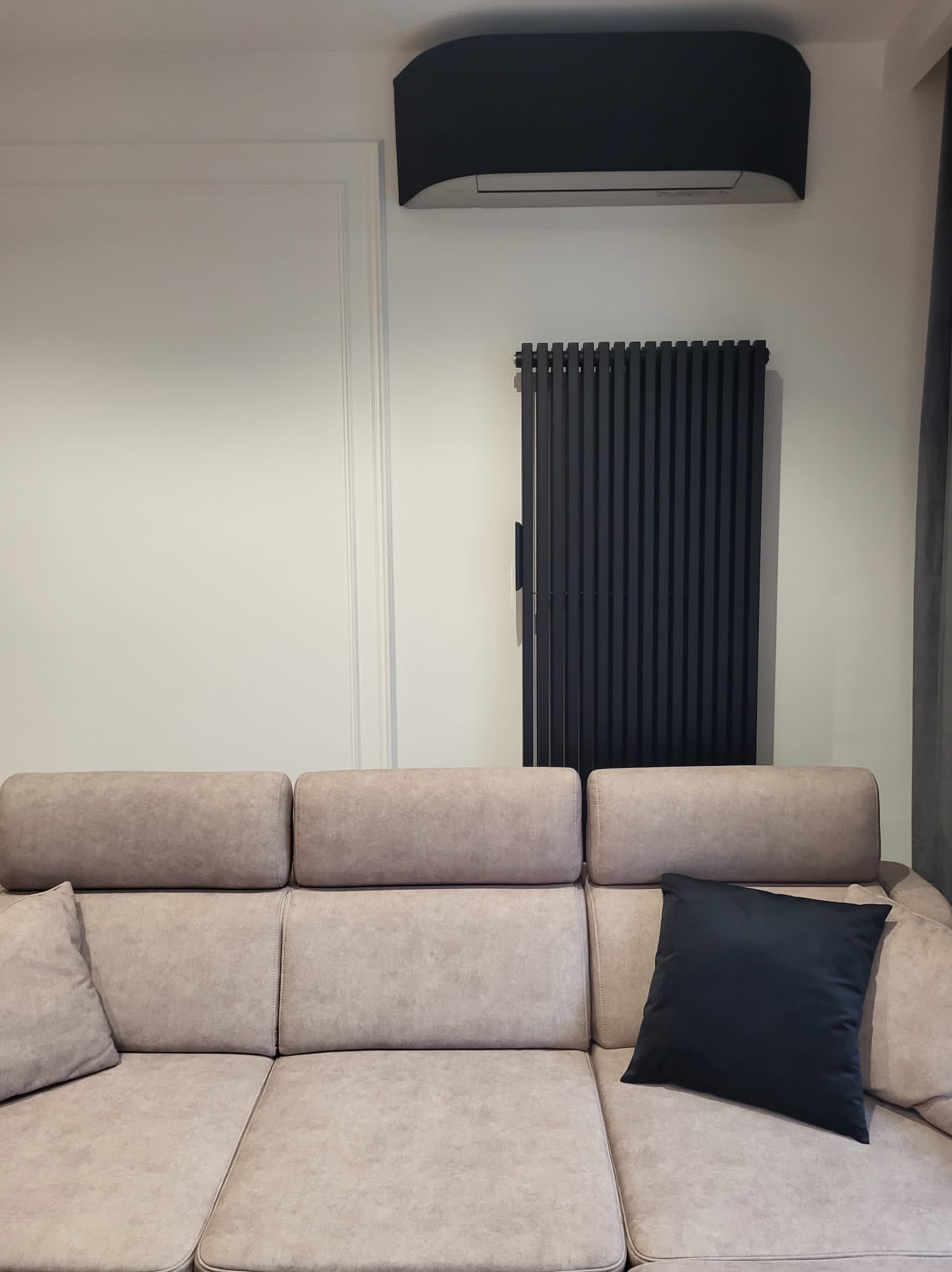 Nowoczesny salon z beżową sofą, czarnym grzejnikiem i klimatyzatorem. Minimalistyczny design, stonowana kolorystyka, eleganckie wykończenie ściany.
