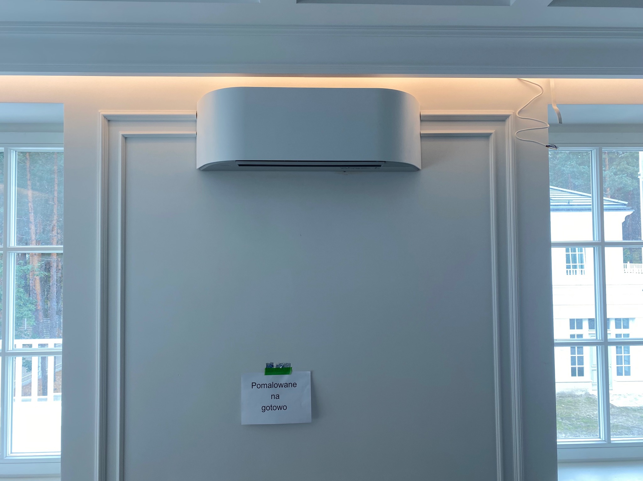 Biały klimatyzator na ścianie z dekoracyjną sztukaterią. Widoczne okna i karteczka z napisem 'Pomalowane na gotowo'. Oświetlenie sufitowe podkreśla nowoczesny design.