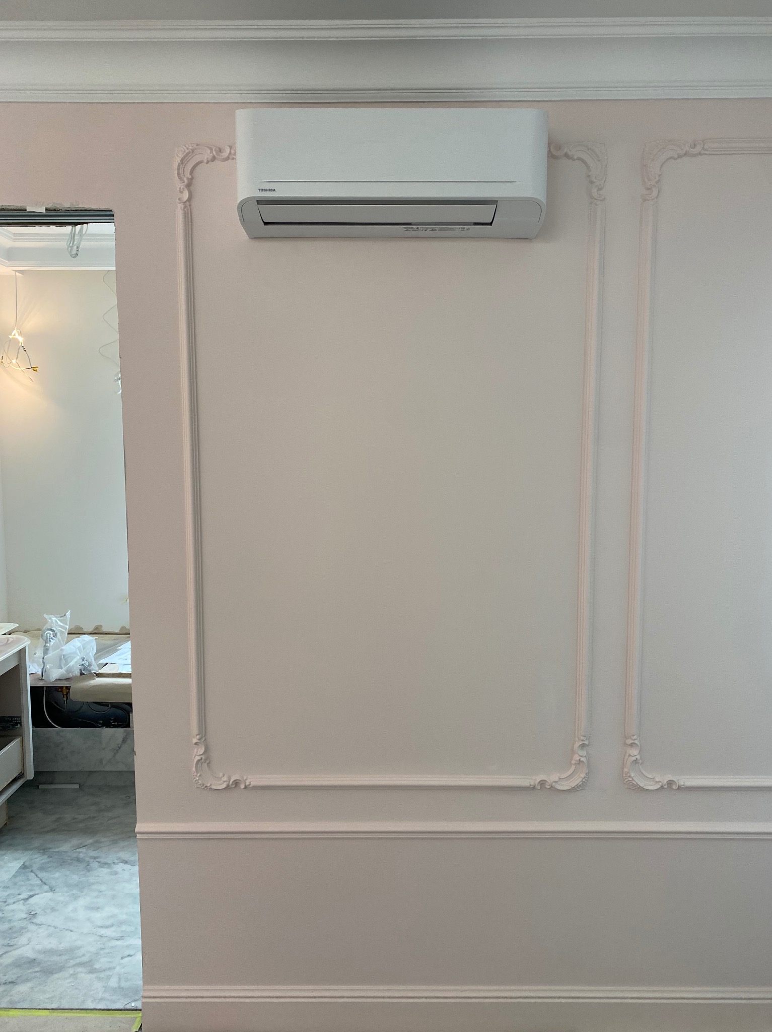 Biały klimatyzator Toshiba zamontowany na ścianie z ozdobnymi listwami w pastelowym odcieniu. Widok na wnętrze przez otwarte drzwi.