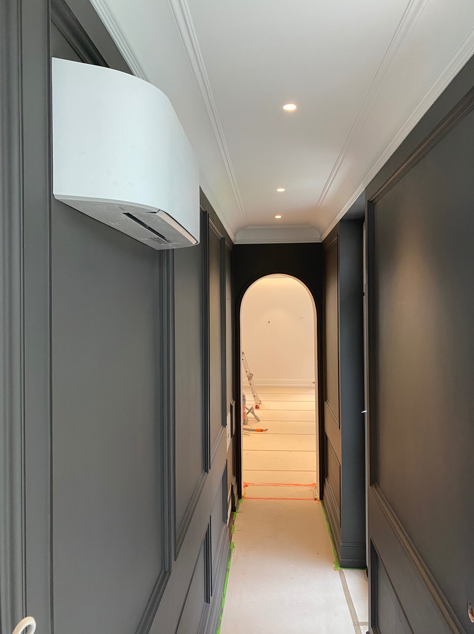 Nowoczesny klimatyzator ścienny w szarym korytarzu z dekoracyjnymi panelami i jasnym oświetleniem punktowym. Widok na jasne pomieszczenie przez łukowe przejście.