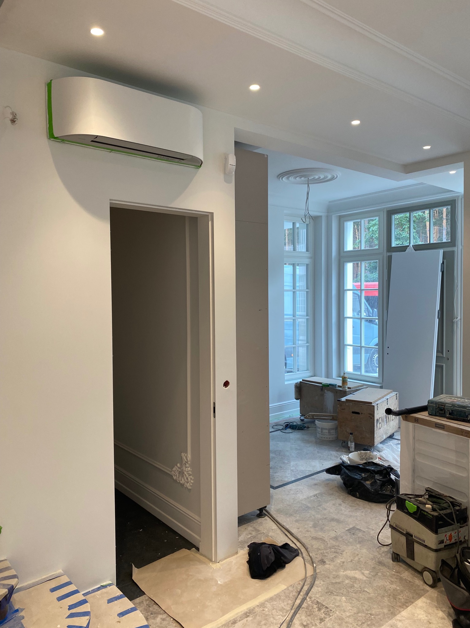 Biały klimatyzator na ścianie w jasnym wnętrzu, zabezpieczony taśmą malarską. Widoczne elementy dekoracyjne sufitu i fragmenty podłogi w trakcie prac wykończeniowych.