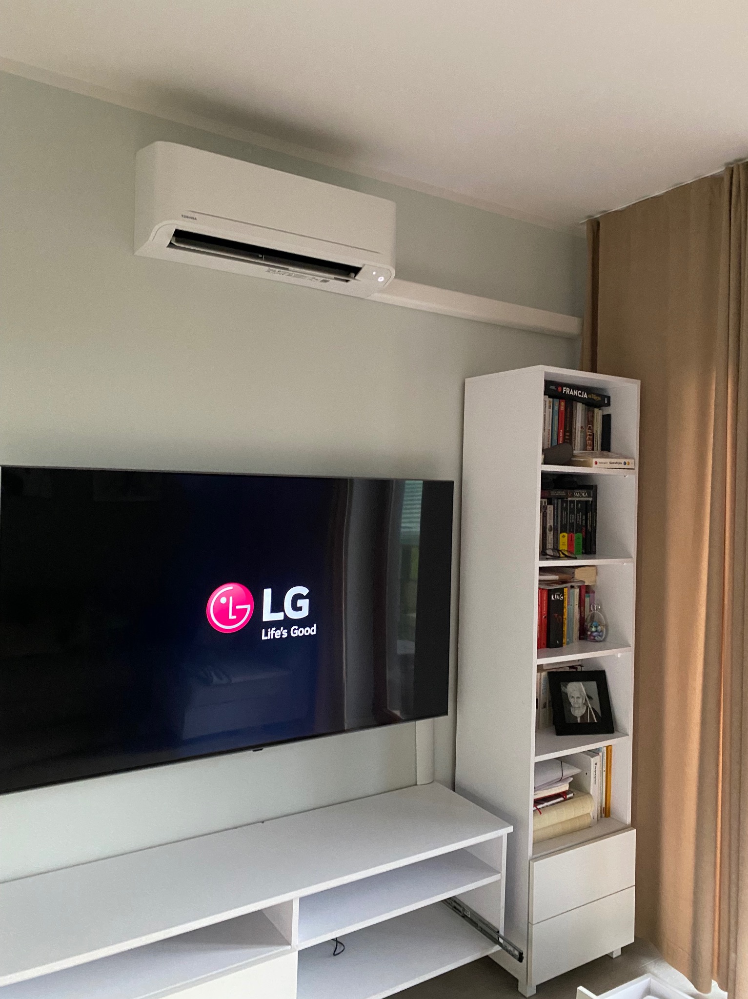 Biały klimatyzator Toshiba zamontowany nad telewizorem LG w nowoczesnym salonie z beżowymi zasłonami i regałem z książkami.