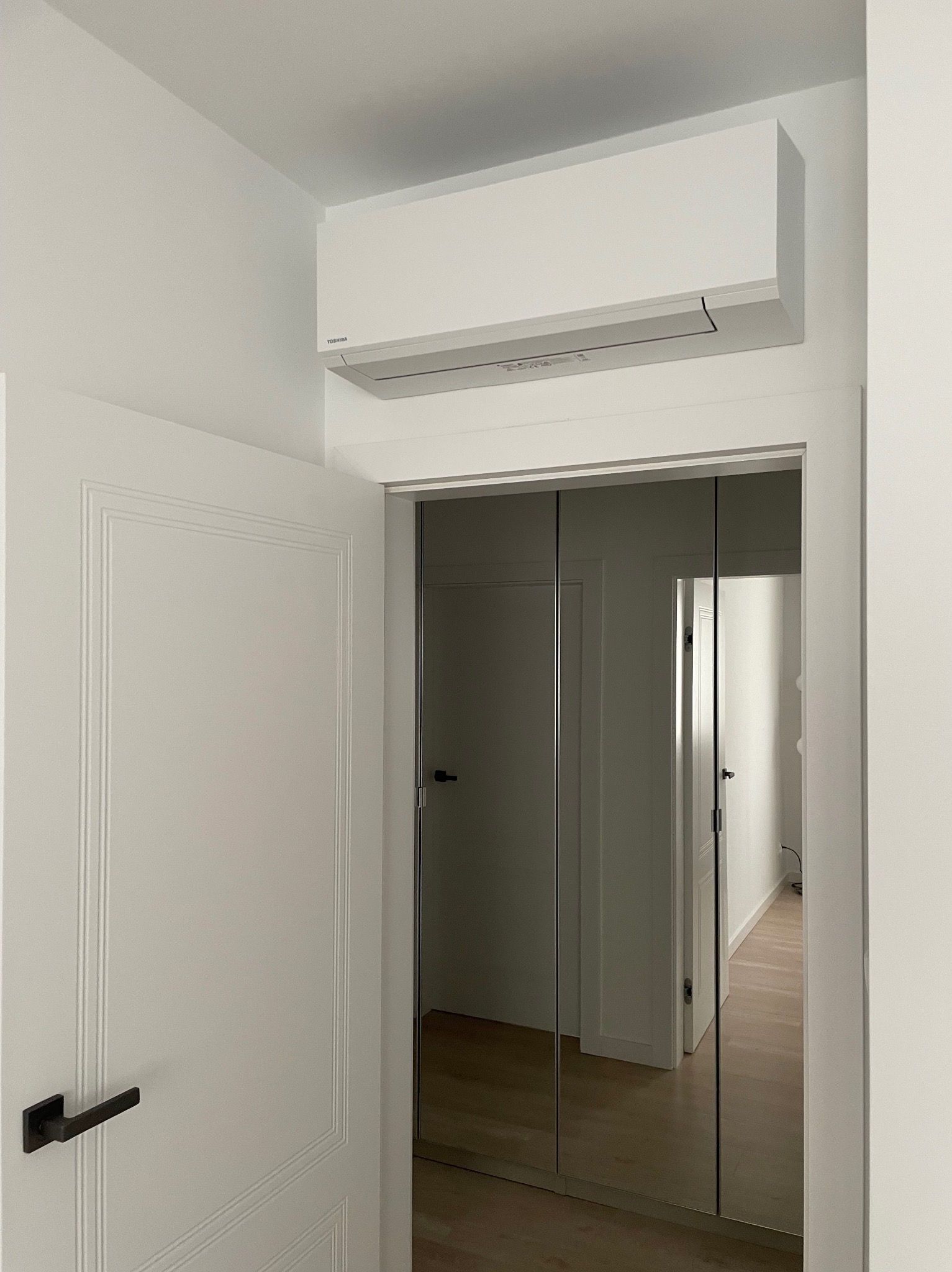 Biała klimatyzacja Toshiba zamontowana nad lustrzanymi drzwiami przesuwnymi w jasnym wnętrzu. Widoczny fragment otwartych drzwi z czarną klamką.