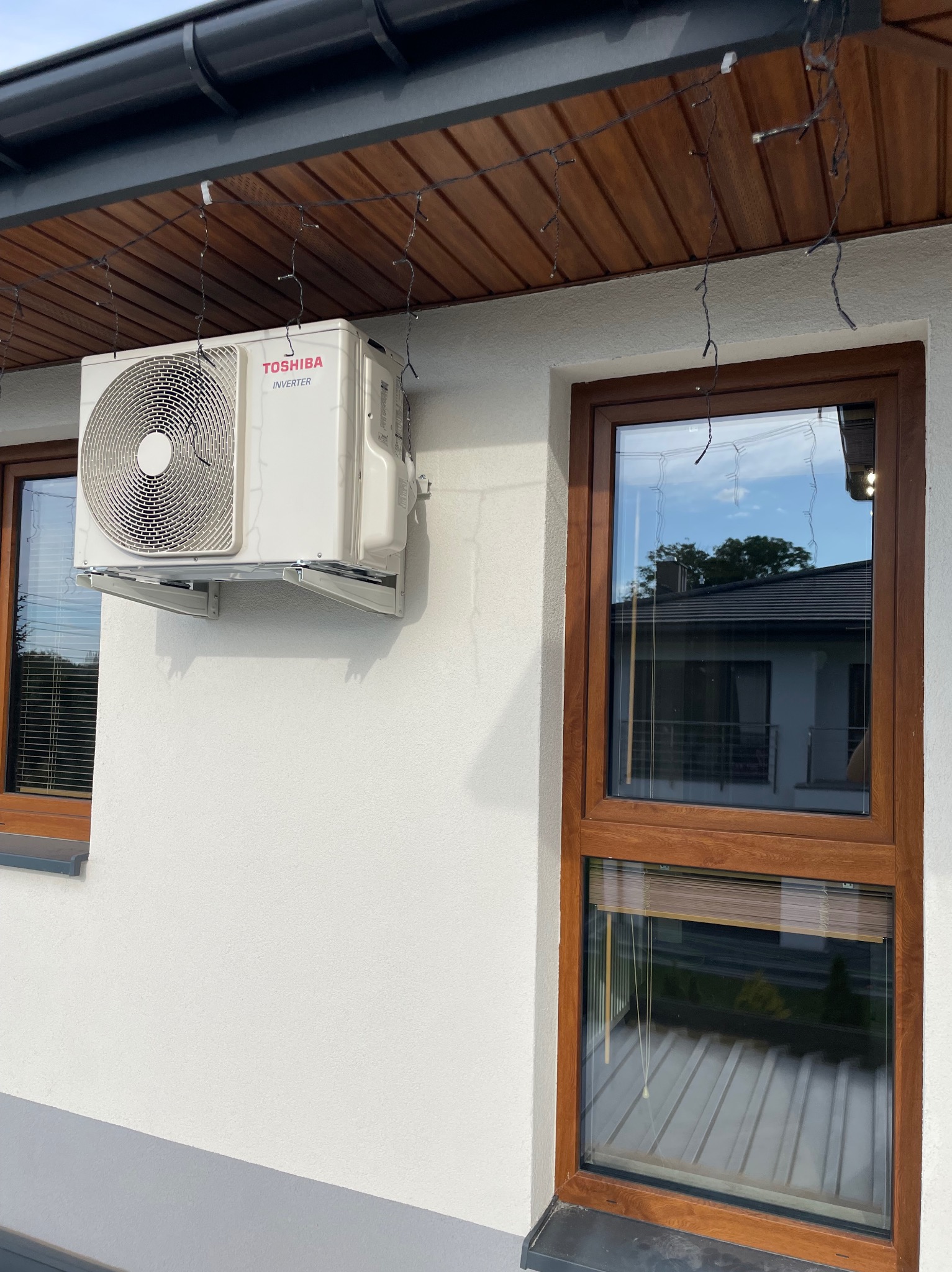 Biała jednostka zewnętrzna klimatyzacji Toshiba zamontowana na ścianie domu, widoczna z boku, z oknami o drewnianych ramach w tle. Dekoracyjne lampki wiszą nad urządzeniem.