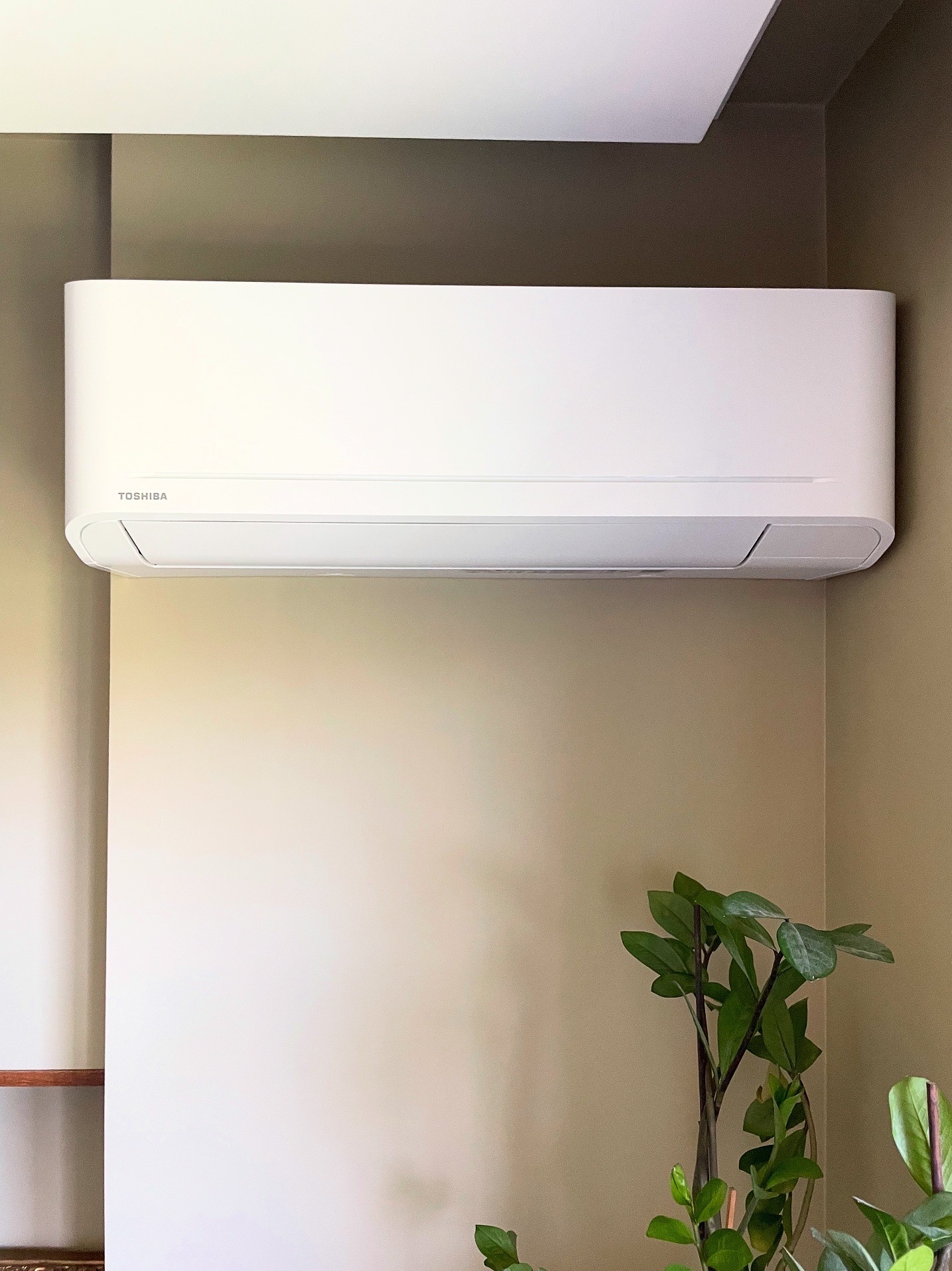 Nowoczesny, biały klimatyzator Toshiba zamontowany na beżowej ścianie w jasnym wnętrzu. Roślina doniczkowa w prawym dolnym rogu dodaje naturalnego akcentu.