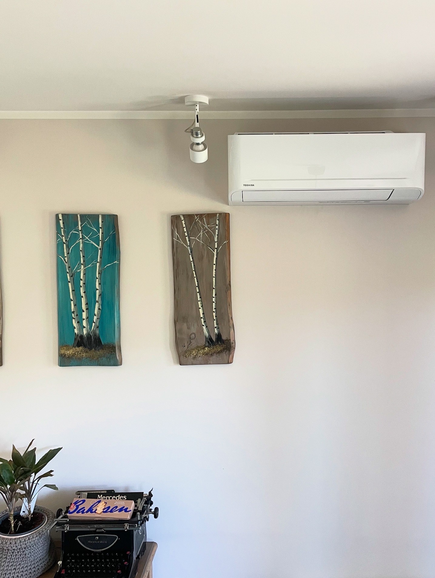 Biała klimatyzacja Toshiba zamontowana na ścianie w Warszawie, nad obrazami z motywem brzozy i starą maszyną do pisania. Widoczna lampa sufitowa.