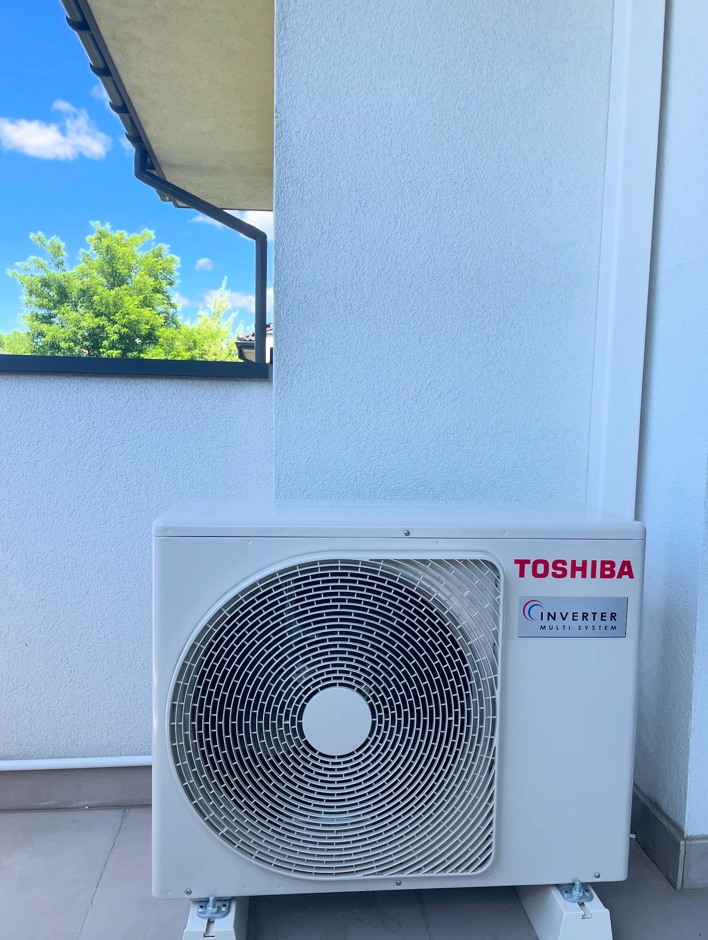 Biała, zewnętrzna jednostka klimatyzacji Toshiba Inverter Multi System zamontowana na jasnej elewacji budynku z widokiem na zieleń za oknem.