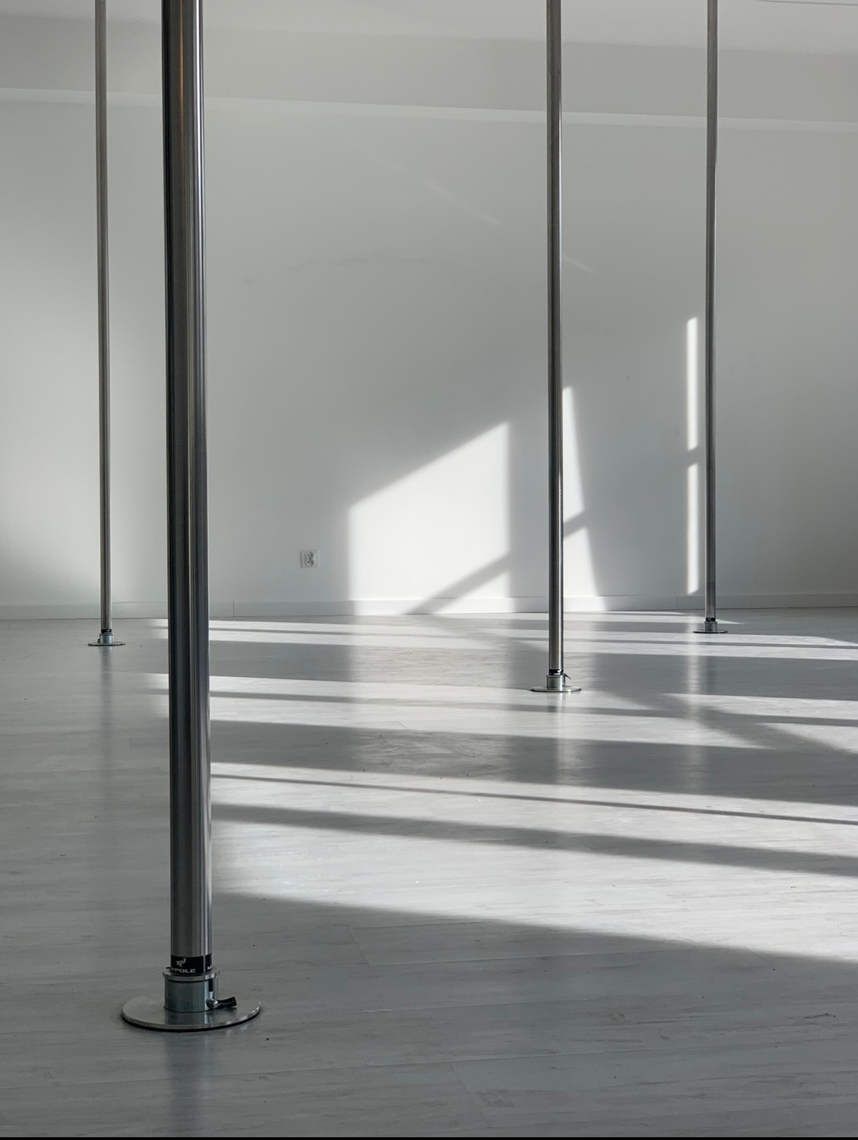 Jasne studio z rzędami srebrnych rur do tańca, oświetlone naturalnym światłem tworzącym geometryczne cienie na podłodze i ścianie. Minimalistyczna przestrzeń.