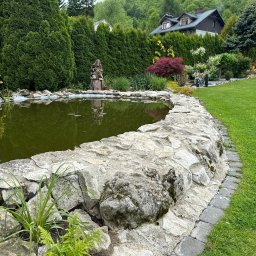 Dawid Piwowarczyk PRODAW - Oczko wodne z kamiennym brzegiem w Jerzmanowicach, otoczone zielenią i wysokimi tujami. W tle dom z ciemnym dachem i biały samochód dostawczy.