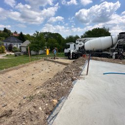 Dawid Piwowarczyk PRODAW - Przygotowanie zbrojenia pod wylewkę fundamentu z użyciem siatki stalowej i dystansów, z betoniarką w tle, na tle zabudowy jednorodzinnej w słoneczny dzień.