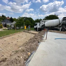 Przygotowanie zbrojenia pod wylewkę fundamentu z użyciem siatki stalowej i dystansów, z betoniarką w tle, na tle zabudowy jednorodzinnej w słoneczny dzień.