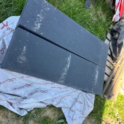 Dawid Piwowarczyk PRODAW - Dwa czarne bloki styropianowe na folii z logo Kost-Bet, leżące na trawie. Widoczne fragmenty obuwia i narzędzi. Przygotowania do izolacji fundamentów.