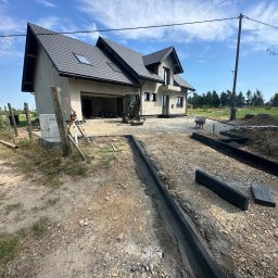 Dawid Piwowarczyk PRODAW - Budowa domu jednorodzinnego: przygotowanie terenu pod kostkę brukową, widoczne obrzeża i mini koparka na żwirowym podłożu, dom w tle.