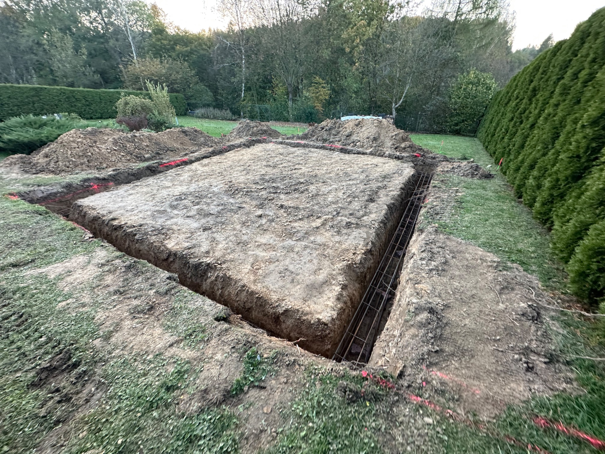 Wykop pod fundament z widocznym zbrojeniem na działce z zielonym trawnikiem i żywopłotem. Czerwone znaczniki wyznaczają obrys. Ziemia usypana wokół wykopu.