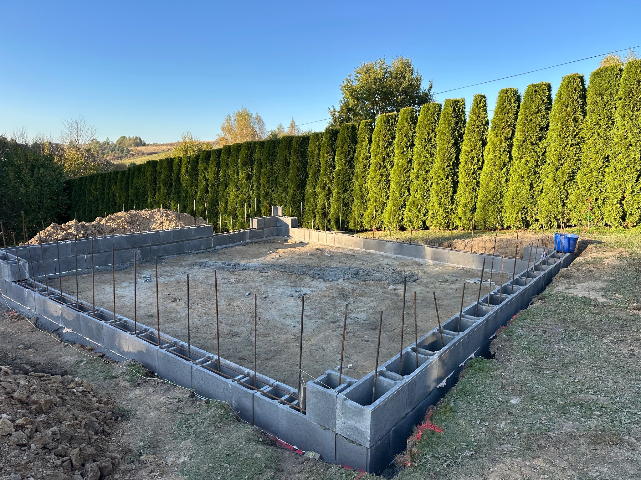 Fundament z bloczków betonowych z wystającymi prętami zbrojeniowymi na tle zielonych tuj i błękitnego nieba, widok z poziomu gruntu.