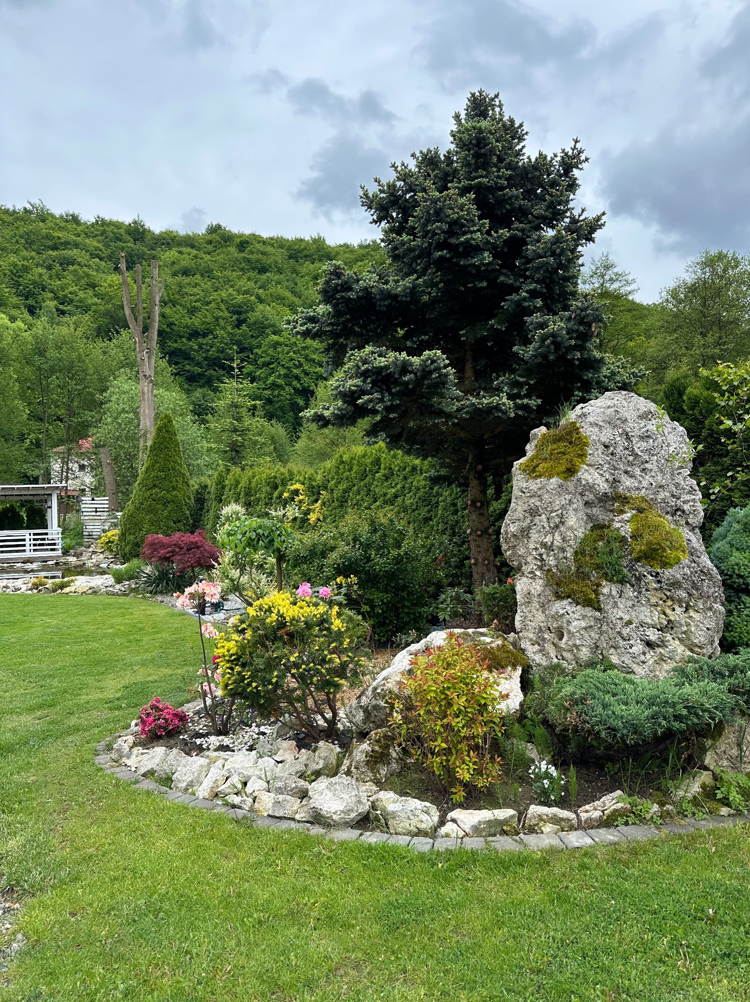 Urokliwy ogród z dużym kamieniem porośniętym mchem, otoczony różnorodną roślinnością i kwiatami. W tle widoczny zielony las i elementy architektury ogrodowej.