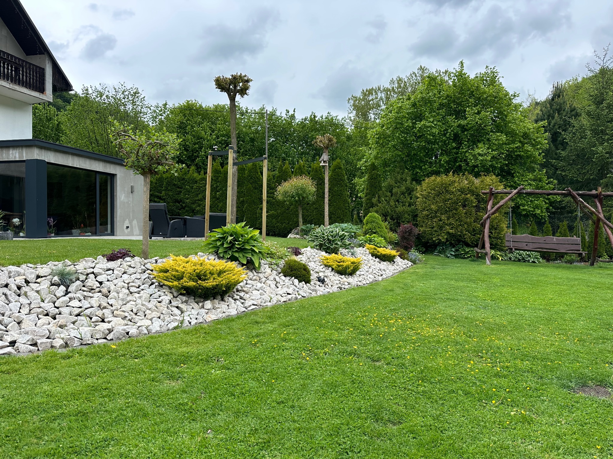 Urokliwy ogród z białymi kamieniami, dekoracyjnymi krzewami i drewnianą huśtawką w tle, tworzący harmonijną przestrzeń przy nowoczesnym domu.