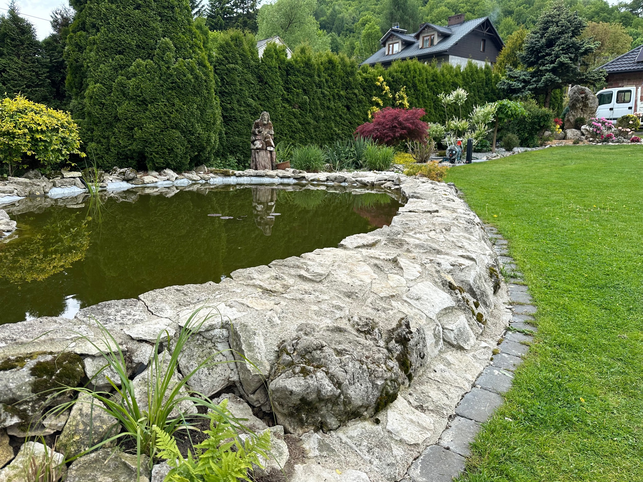 Oczko wodne z kamiennym brzegiem w Jerzmanowicach, otoczone zielenią i wysokimi tujami. W tle dom z ciemnym dachem i biały samochód dostawczy.