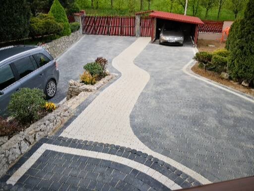 Ułożona kostka brukowa w odcieniach szarości i bieli, tworząca wzór ścieżki do garażu. Widoczne rośliny wzdłuż krawędzi i zaparkowany samochód.