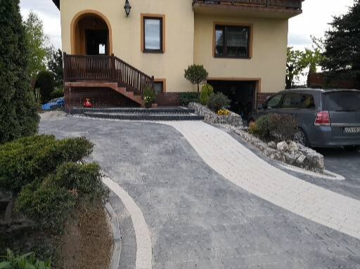 Ułożona kostka brukowa tworzy podjazd do domu z garażem. Jasne obrzeża podkreślają kształt. Widoczny fragment domu z drewnianymi schodami i zaparkowany samochód.