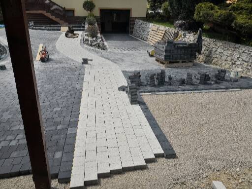 Układanie jasnoszarej kostki brukowej na żwirowym podłożu, widoczne palety z kostką i ukończony fragment ścieżki z ciemnoszarej kostki. Prace w toku.